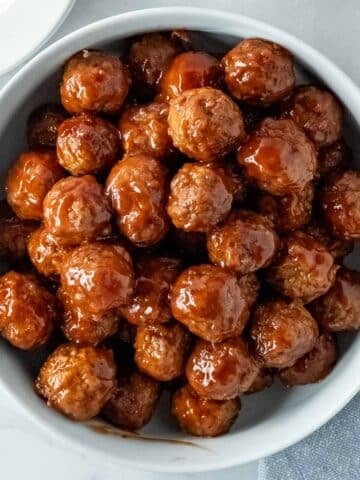 A white bowl filled with grape jelly and ketchup meatballs on a white marble countertop.