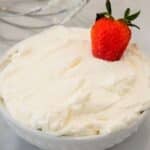 A bowl of whipped cream topped with a single fresh strawberry, with metal beaters visible in the background.