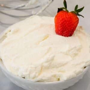 A bowl of whipped cream topped with a single fresh strawberry, with metal beaters visible in the background.