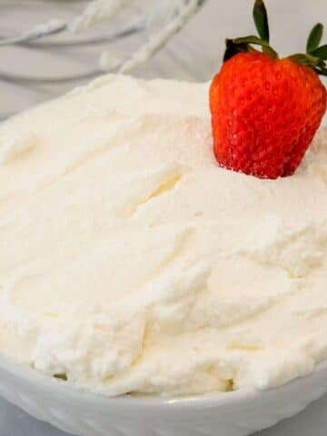 A bowl of whipped cream topped with a single fresh strawberry, with metal beaters visible in the background.