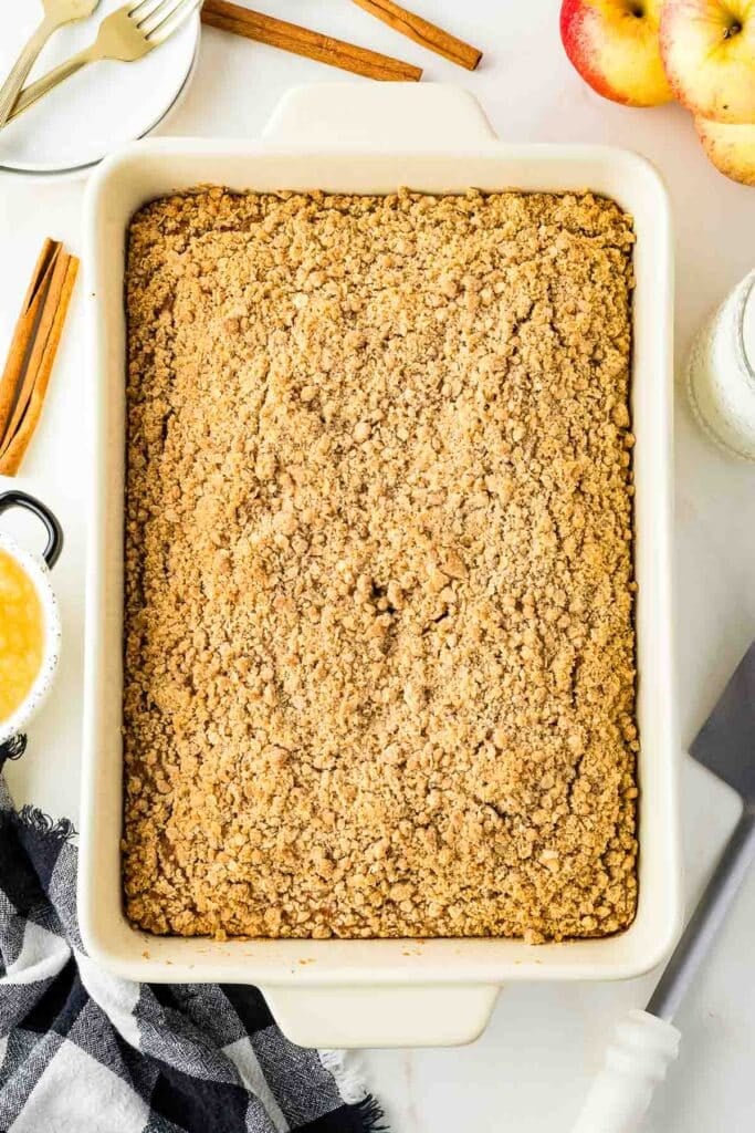 A rectangular baking dish filled with golden-brown Applesauce Crumb Cake on a white surface, surrounded by apples, cinnamon sticks, a black-and-white napkin, and utensils.