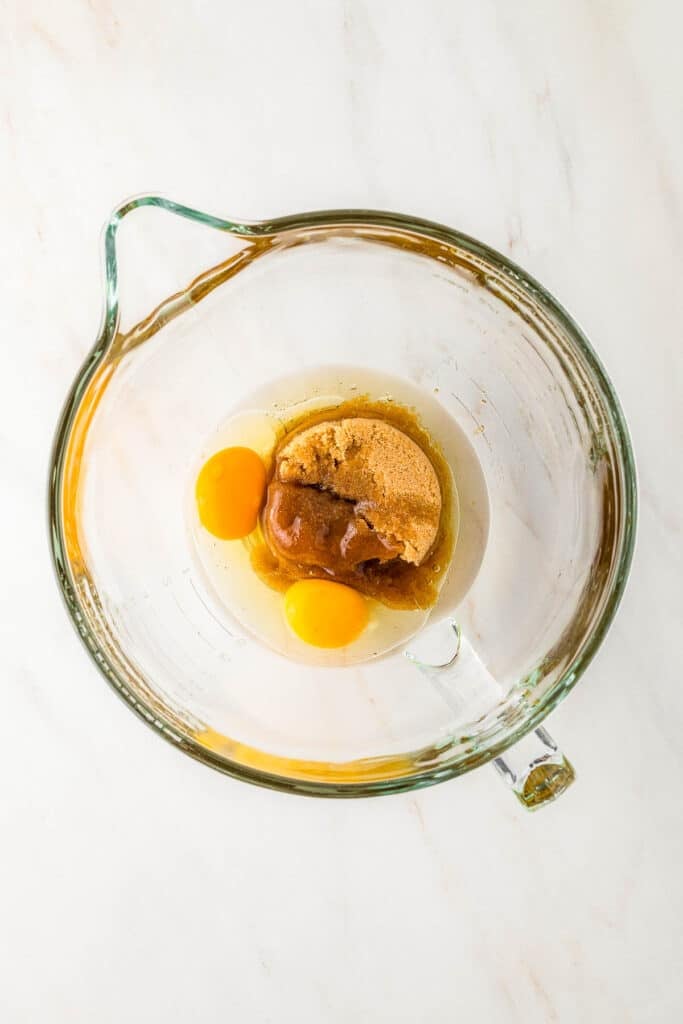 A glass mixing bowl containing two raw eggs, brown sugar, and vanilla extract on a white surface.