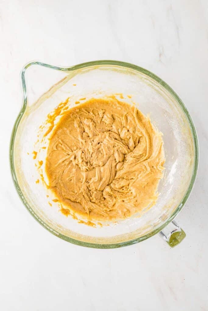 A glass mixing bowl containing partially mixed dough sits on a white marble surface.