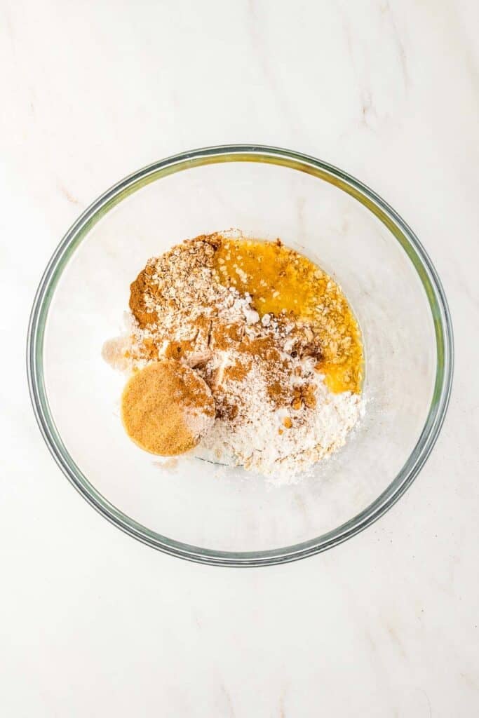 A glass bowl containing flour, sugar, brown sugar, cinnamon, and melted butter on a white surface.