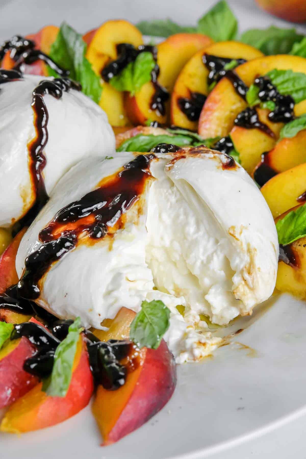 Sliced peaches and whole burrata cheese topped with balsamic glaze and fresh basil leaves on a white plate.