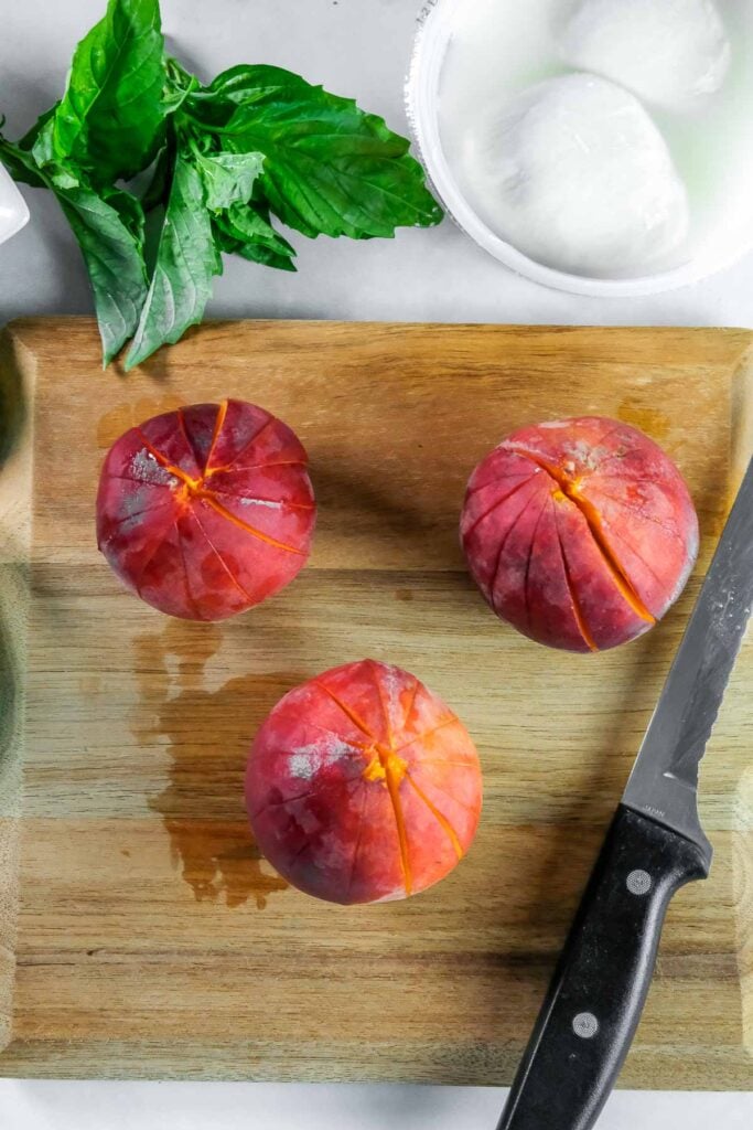 Three peaches with cuts on top are on a wooden cutting board beside a knife and fresh basil leaves.