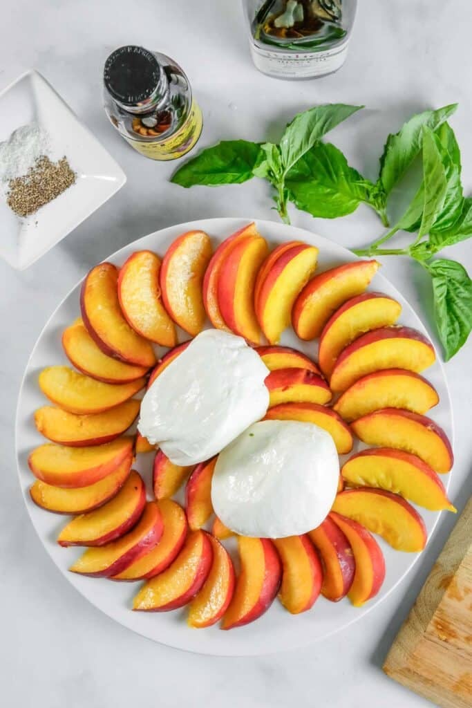 Sliced peaches and two burrata balls arranged on a white plate, with basil leaves, seasoning, and bottles of oil nearby on a marble surface.