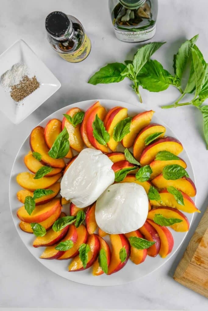 A plate with sliced peaches, two balls of burrata cheese, and fresh basil leaves, with bottles of seasoning, olive oil, and a small dish of salt and pepper nearby.