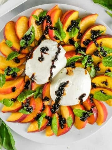 A plate with sliced peaches, fresh burrata, basil leaves, and balsamic glaze.