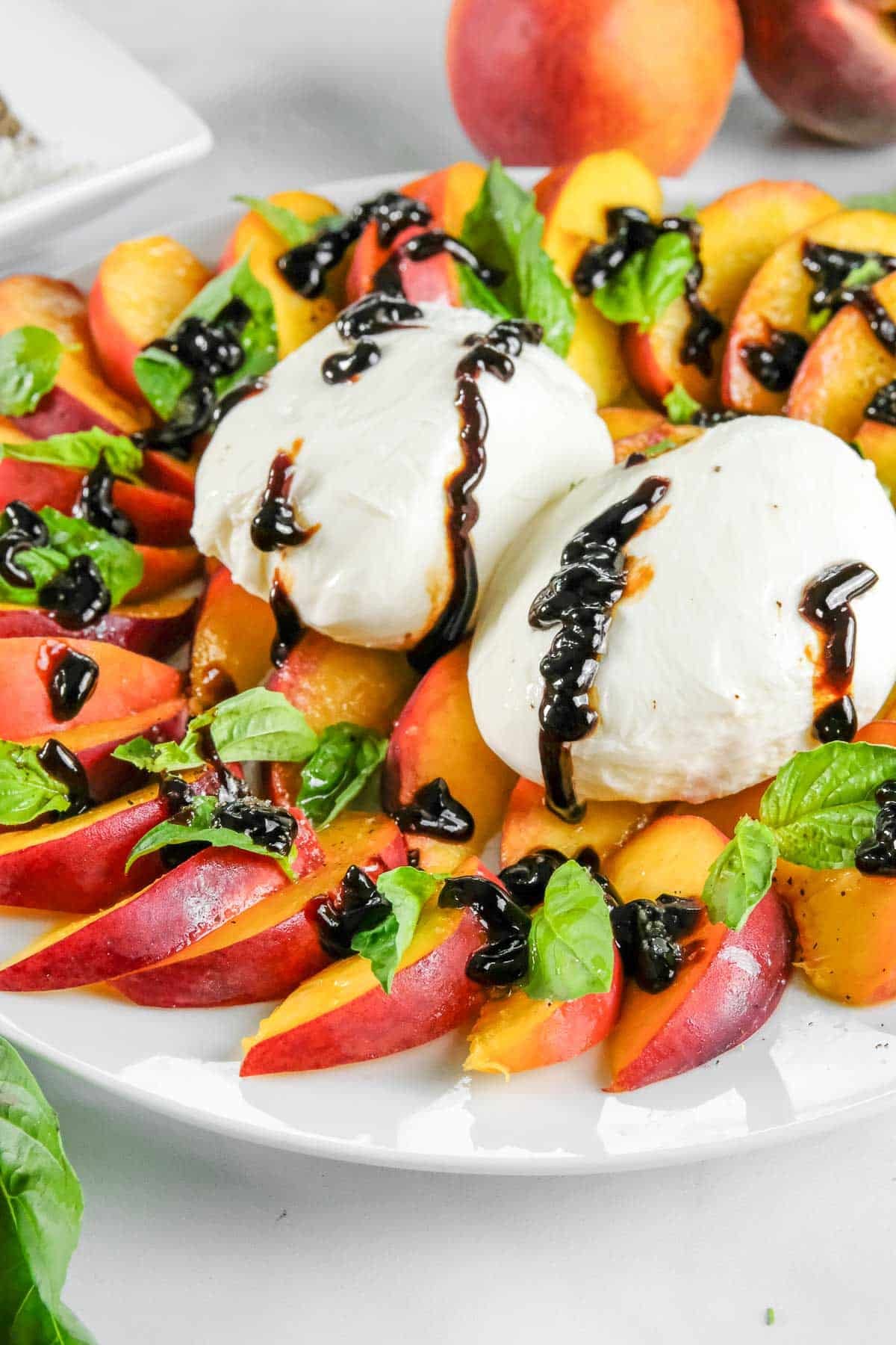 A plate with sliced peaches, fresh basil leaves, two balls of burrata cheese, and a drizzle of balsamic glaze.