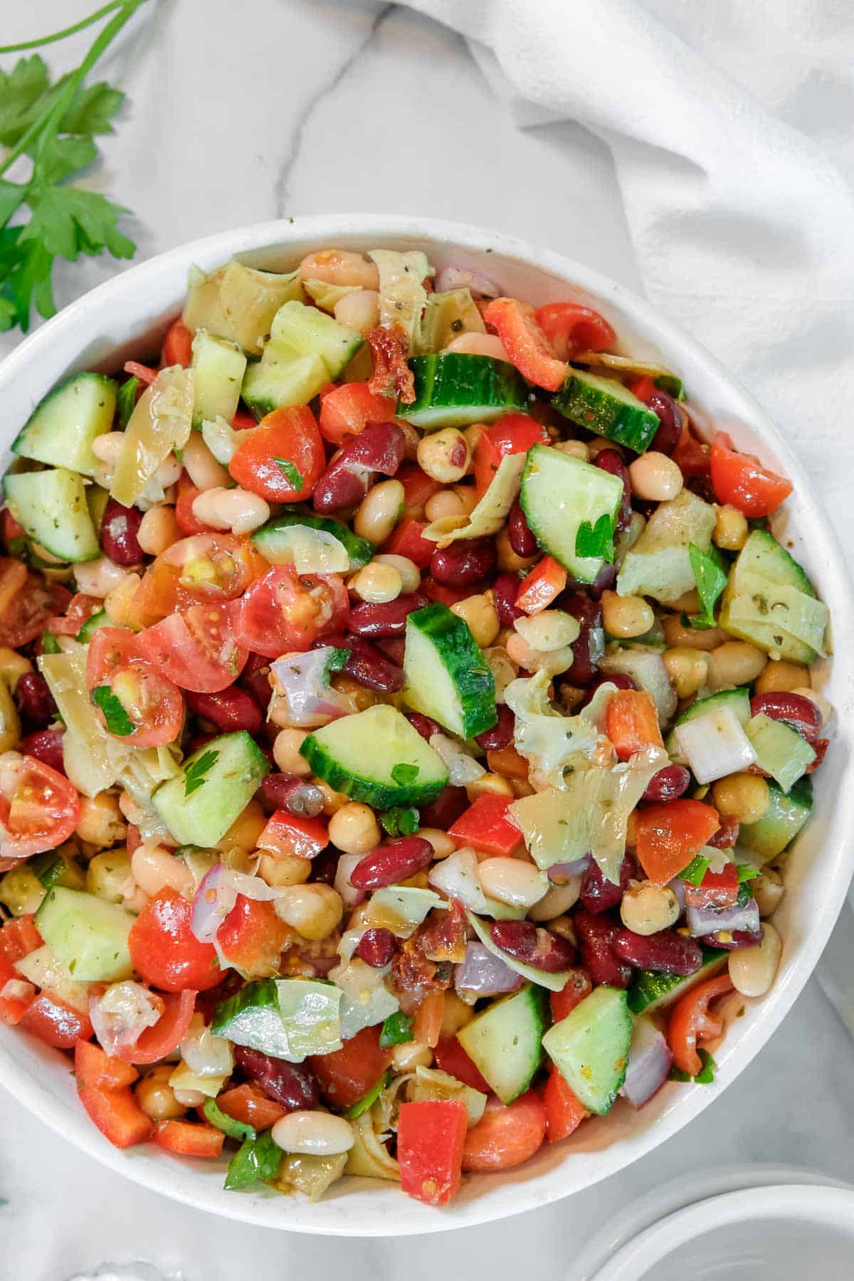 A bowl of dense bean salad with chopped cucumbers, tomatoes, red onions, and parsley on a marble surface.