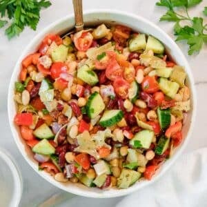 A bowl of dense bean salad with cucumbers, tomatoes, red onions, and red bell peppers, garnished with parsley, on a white surface.