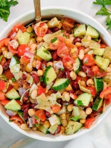 A bowl of dense bean salad with cucumbers, tomatoes, red onions, and red bell peppers, garnished with parsley, on a white surface.