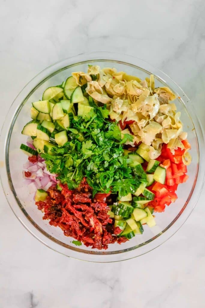 A glass bowl containing chopped cucumber, red bell pepper, red onion, sun-dried tomatoes, artichoke hearts, and fresh parsley arranged in separate sections.