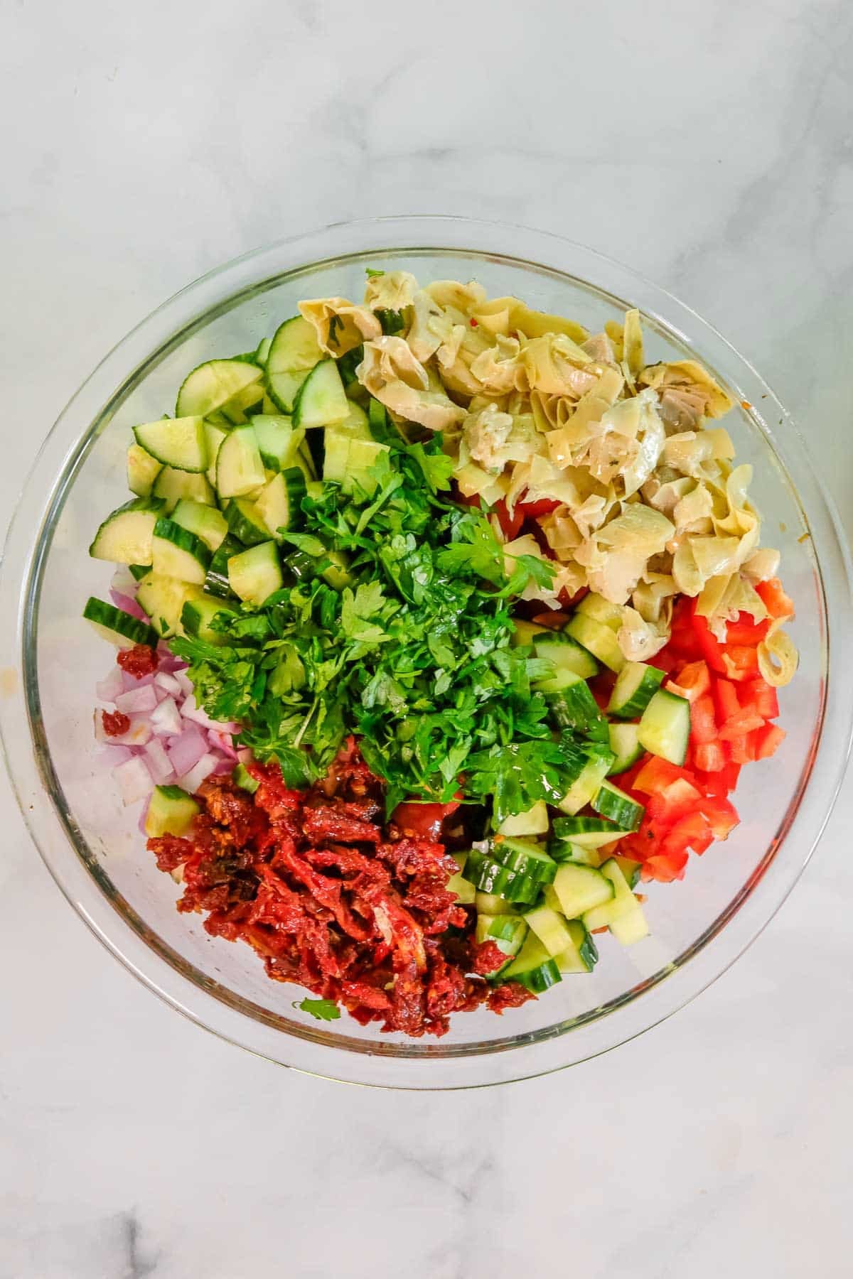 A glass bowl containing chopped cucumber, red bell pepper, red onion, sun-dried tomatoes, artichoke hearts, and fresh parsley arranged in separate sections.