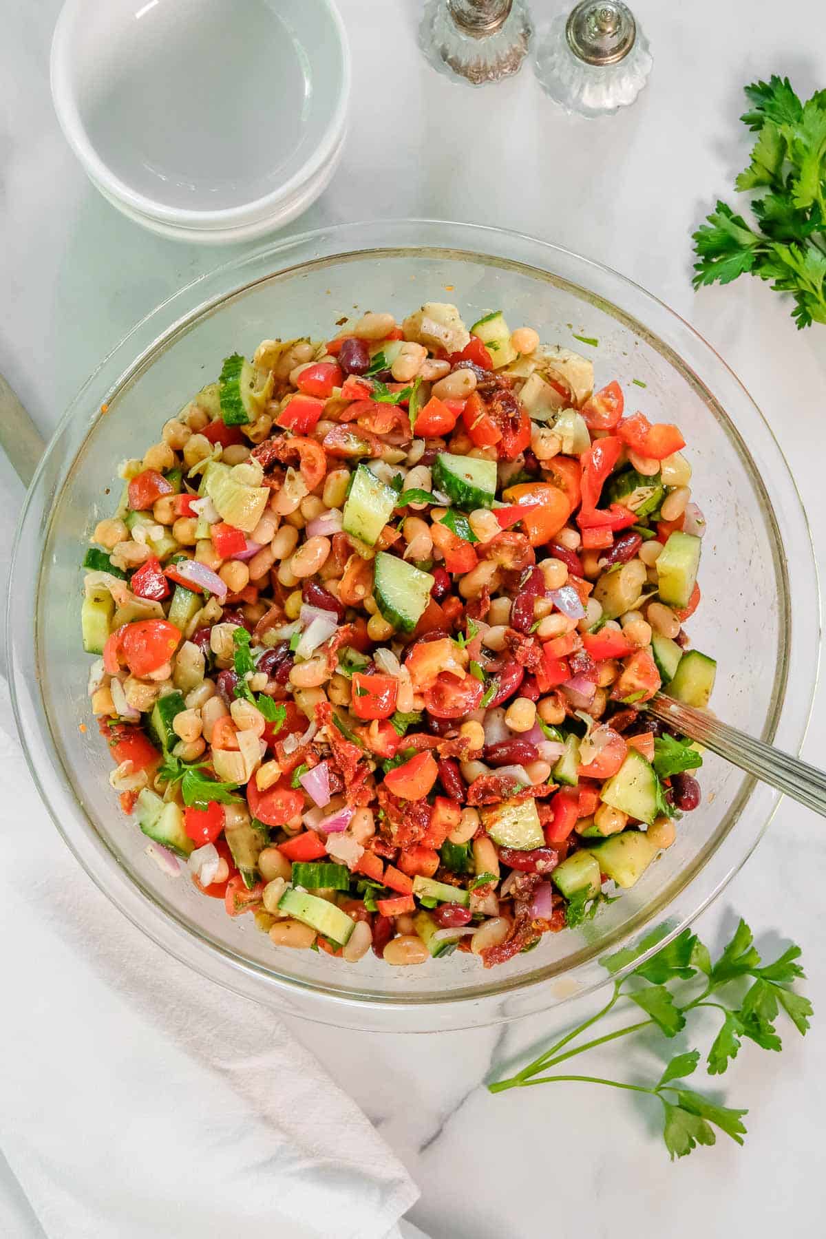 A bowl of mixed dense bean salad with chopped tomatoes, cucumbers, red onions, and herbs, placed on a marble surface with a spoon, parsley, and empty bowls nearby.
