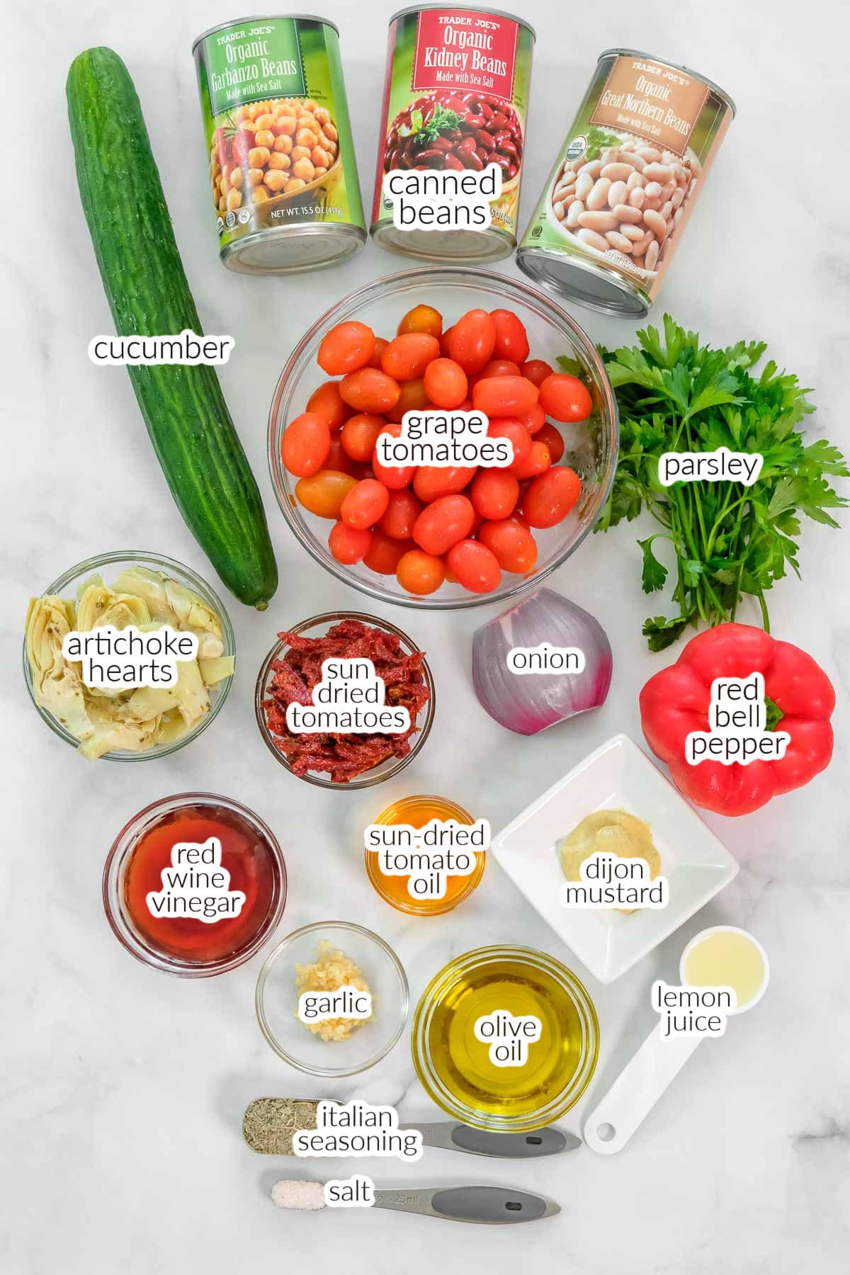 Ingredients for dense bean salad, including canned beans, cucumber, grape tomatoes, parsley, artichoke hearts, sun-dried tomatoes, red bell pepper, onion, and various seasonings and oils.