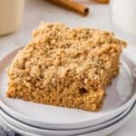A square piece of applesauce crumb cake on a white plate, with a cinnamon stick and glass in the background.