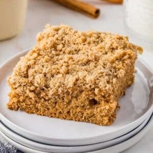 A square piece of applesauce crumb cake on a white plate, with a cinnamon stick and glass in the background.