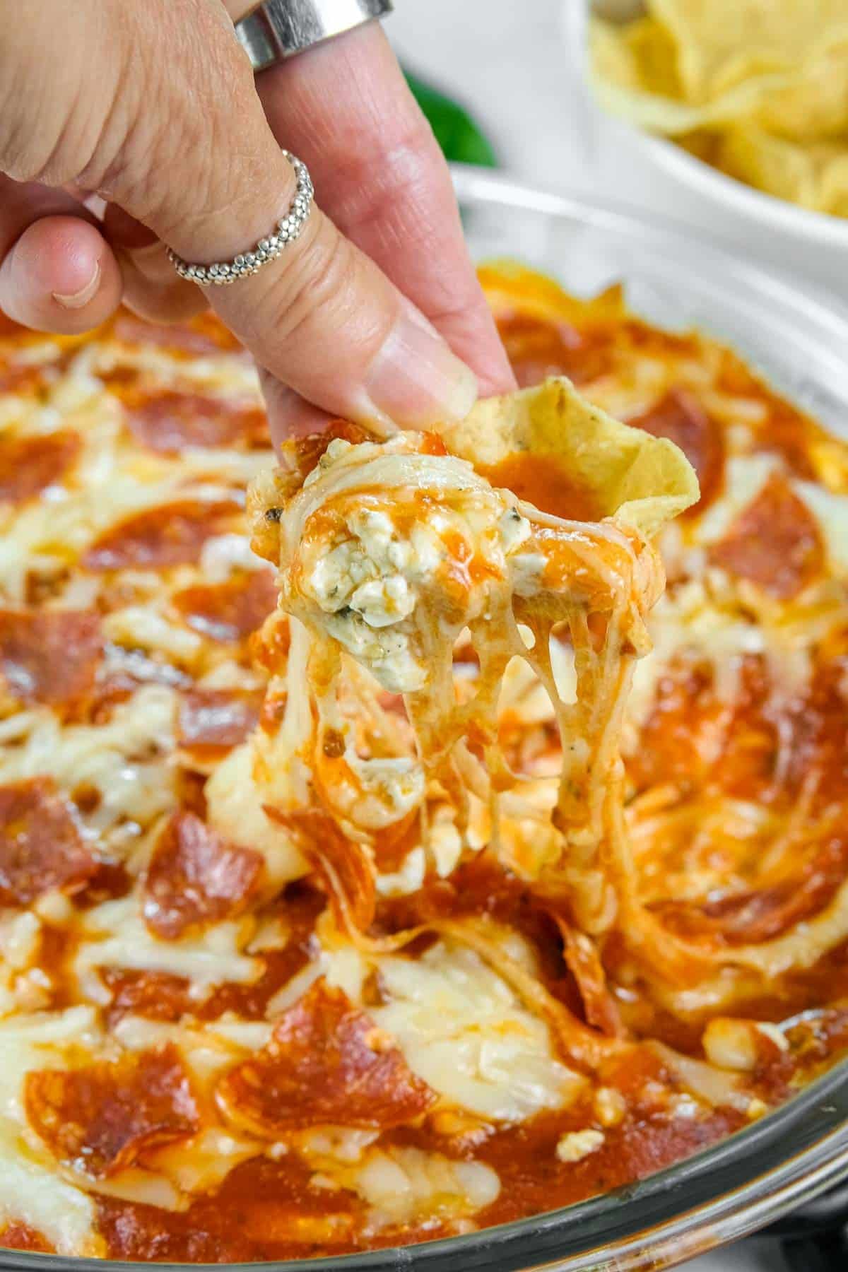 A hand holding a chip scooping cheesy pizza dip from a glass dish, with melted cheese stretching between the dip and the chip.
