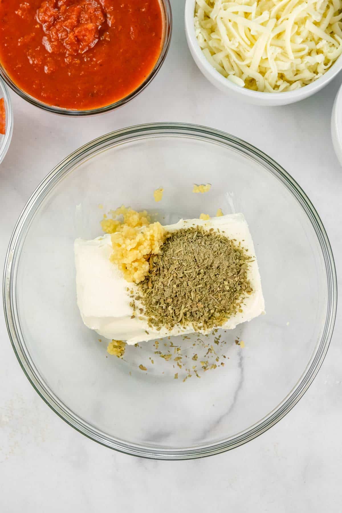 A glass bowl containing cream cheese, minced garlic, and italian seasining.