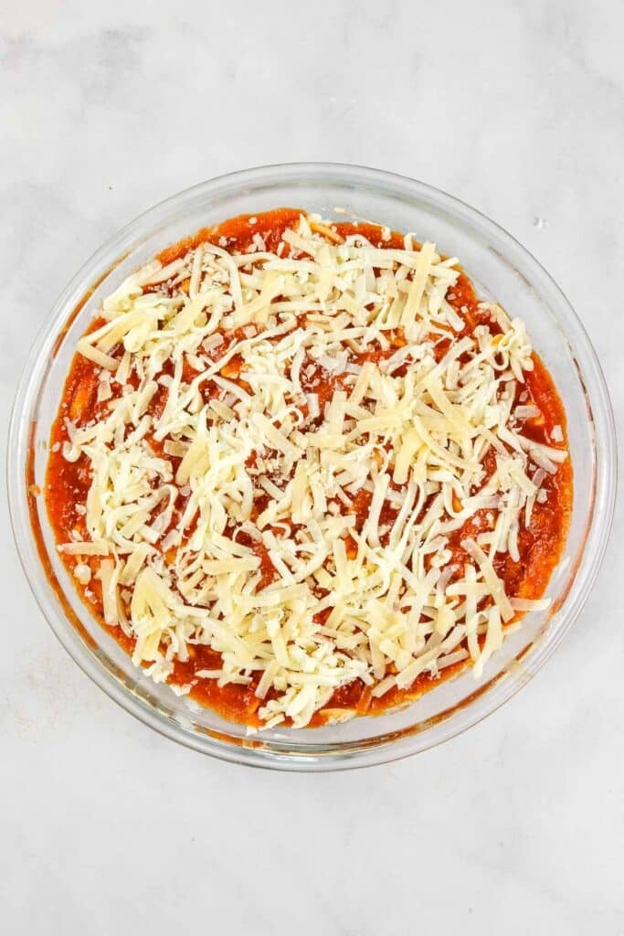 A glass dish filled with a layer of red sauce topped with shredded cheese.