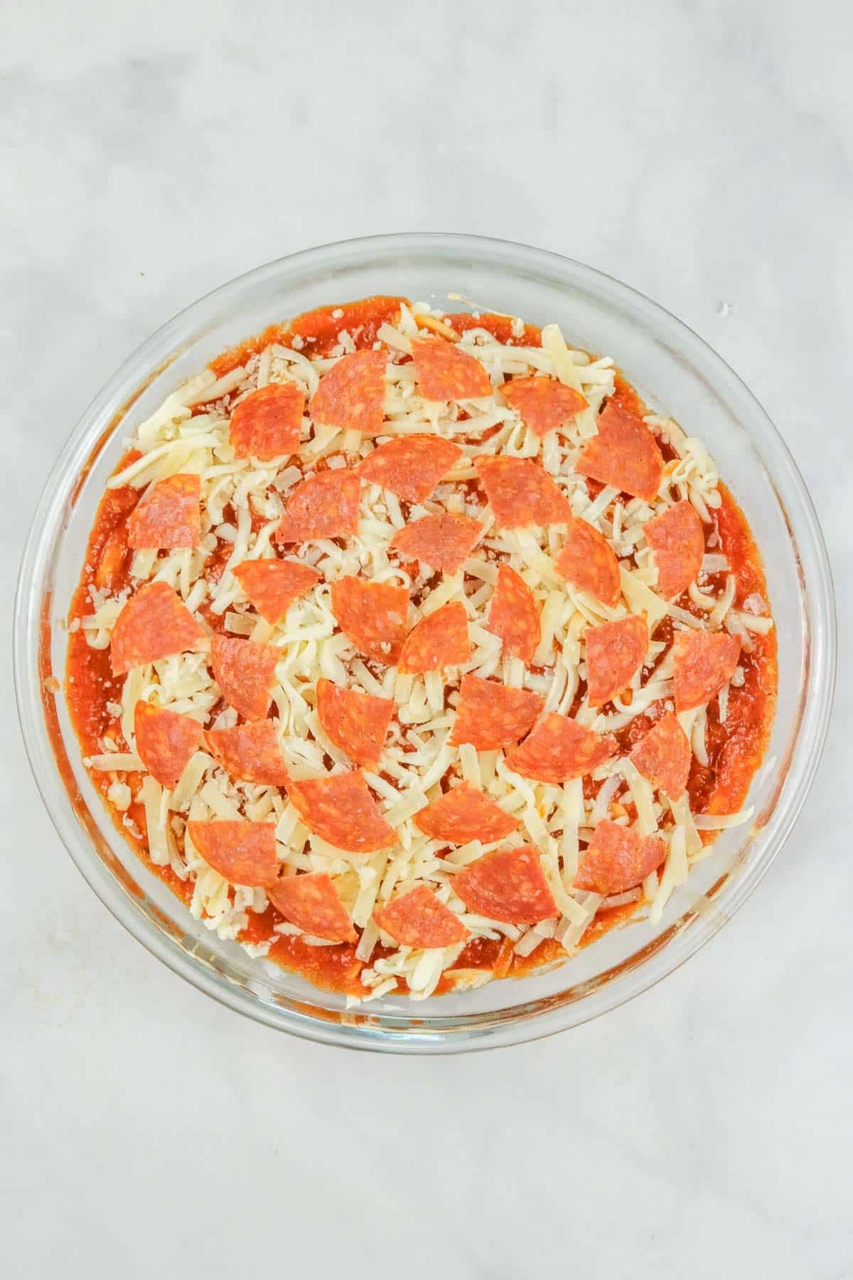 A round glass dish filled with shredded cheese, tomato sauce, and slices of pepperoni, arranged in layers.