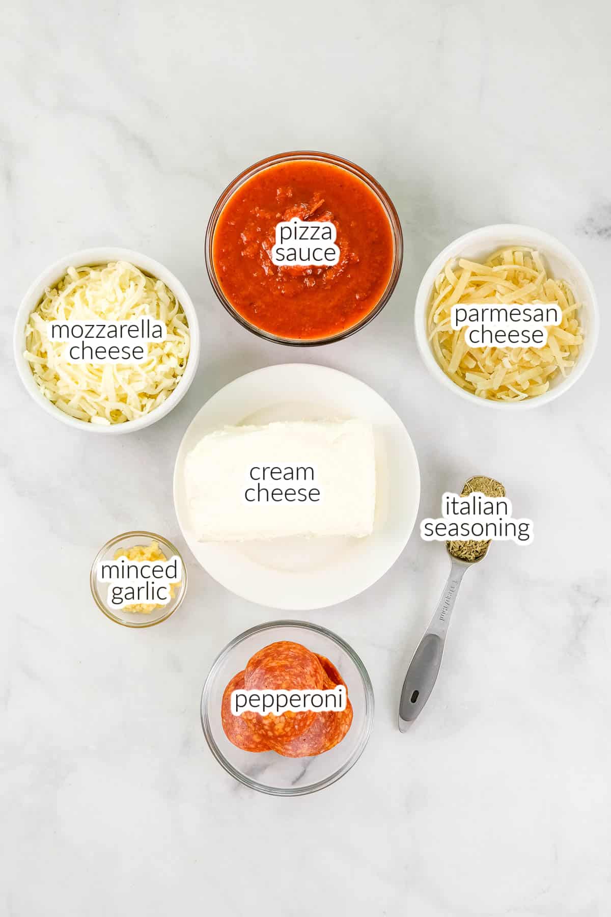 Ingredients for pizza dip including a bowl of pizza sauce, mozzarella cheese, parmesan cheese, minced garlic, pepperoni, cream cheese, and a spoon of Italian seasoning arranged on a marble surface.