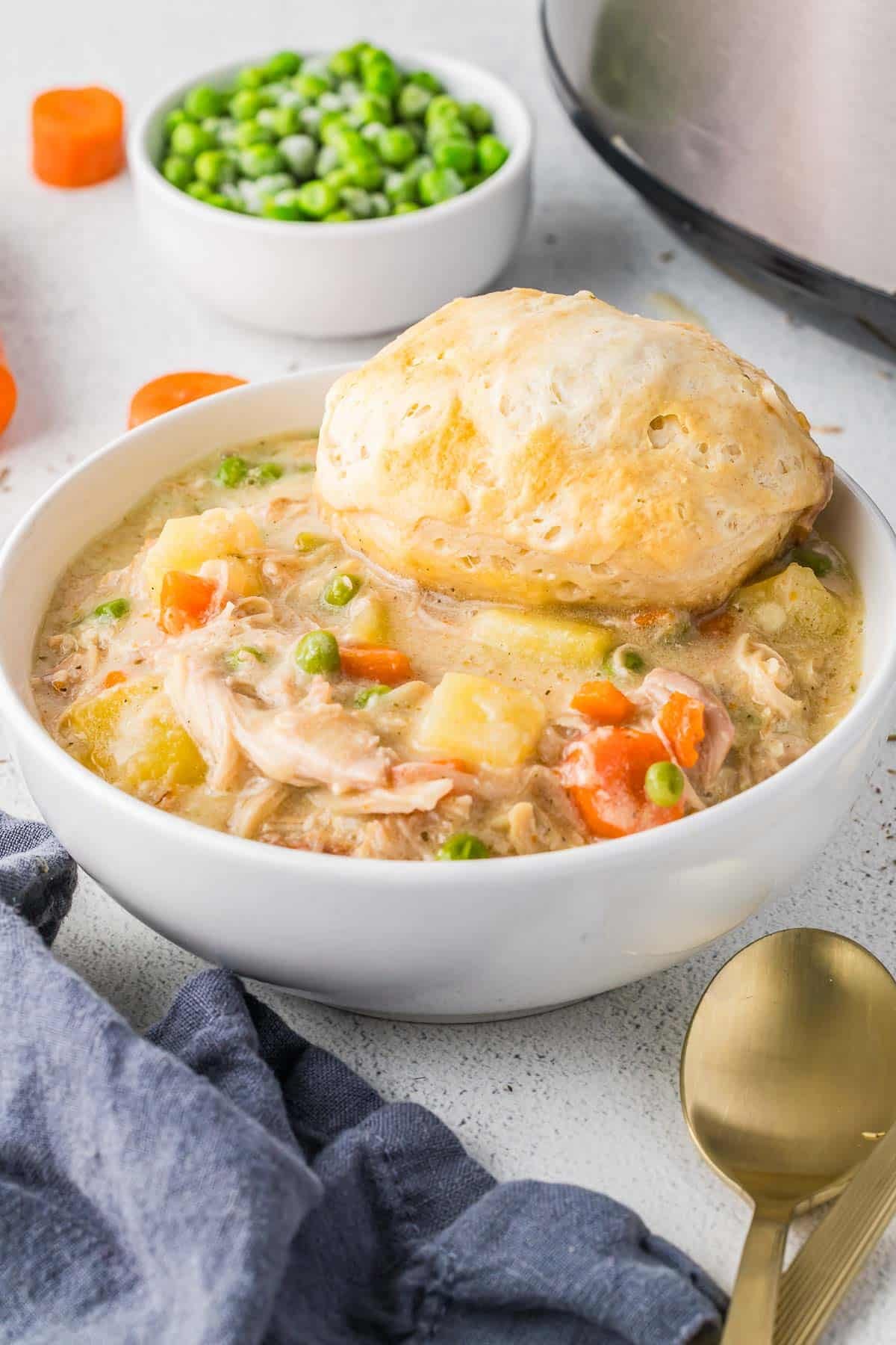 A bowl of slow cooker chicken pot pie with vegetables and a biscuit on top, next to a bowl of peas and a gold spoon on a white surface.
