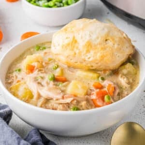 A bowl of slow cooker chicken pot pie with vegetables and a biscuit on top, next to a bowl of peas and a gold spoon on a white surface.