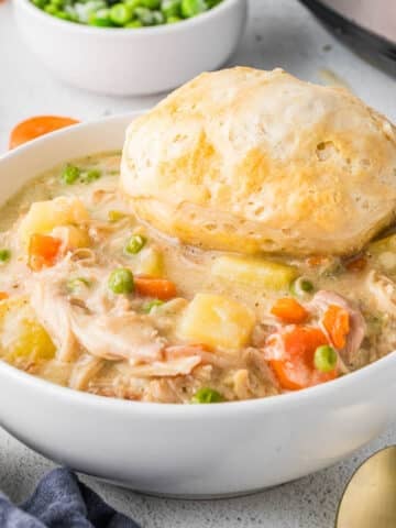 A bowl of slow cooker chicken pot pie with vegetables and a biscuit on top, next to a bowl of peas and a gold spoon on a white surface.