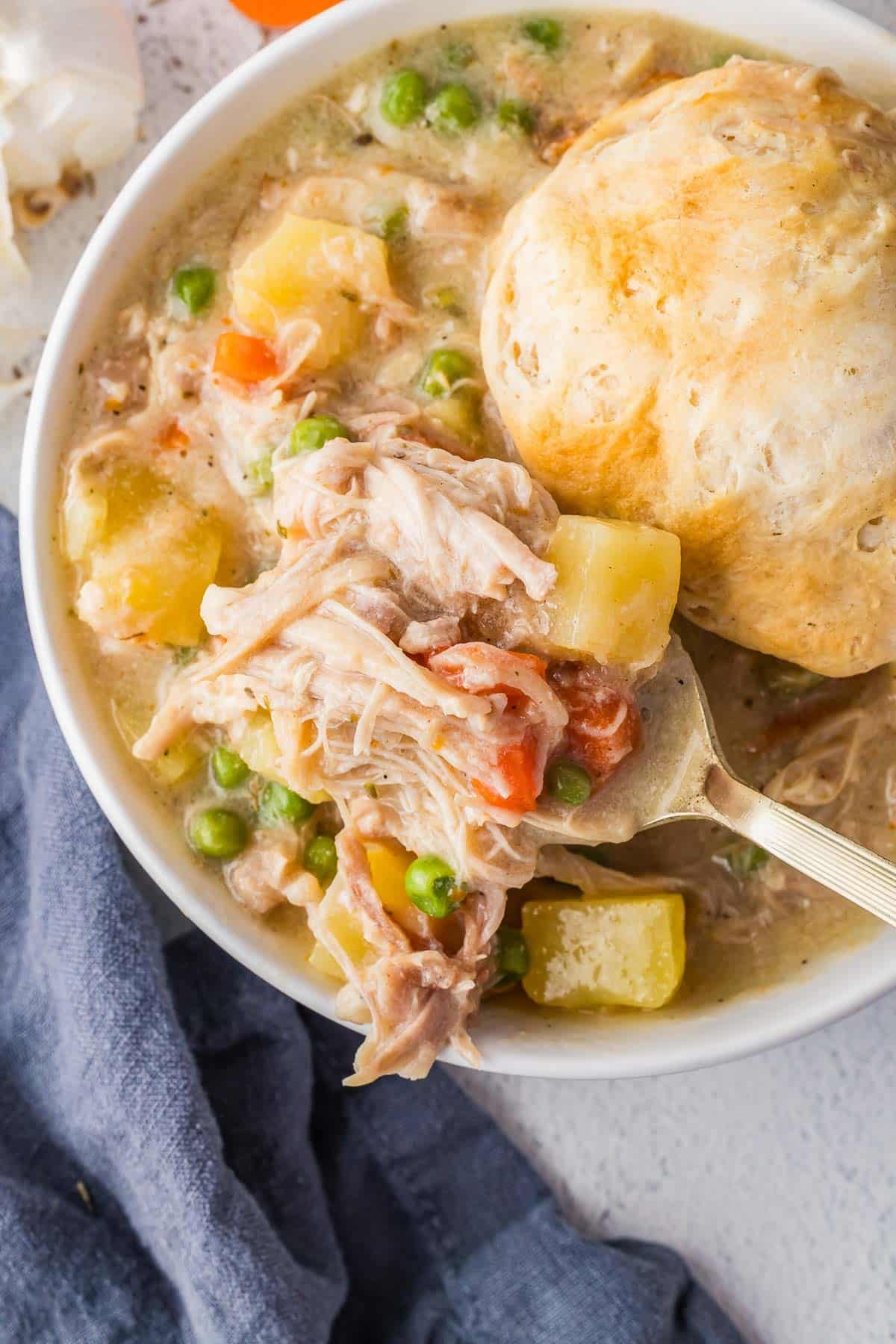 Bowl of chicken pot pie with peas, carrots, potatoes, and a biscuit on top. A spoon is lifting a portion of the stew from the bowl.