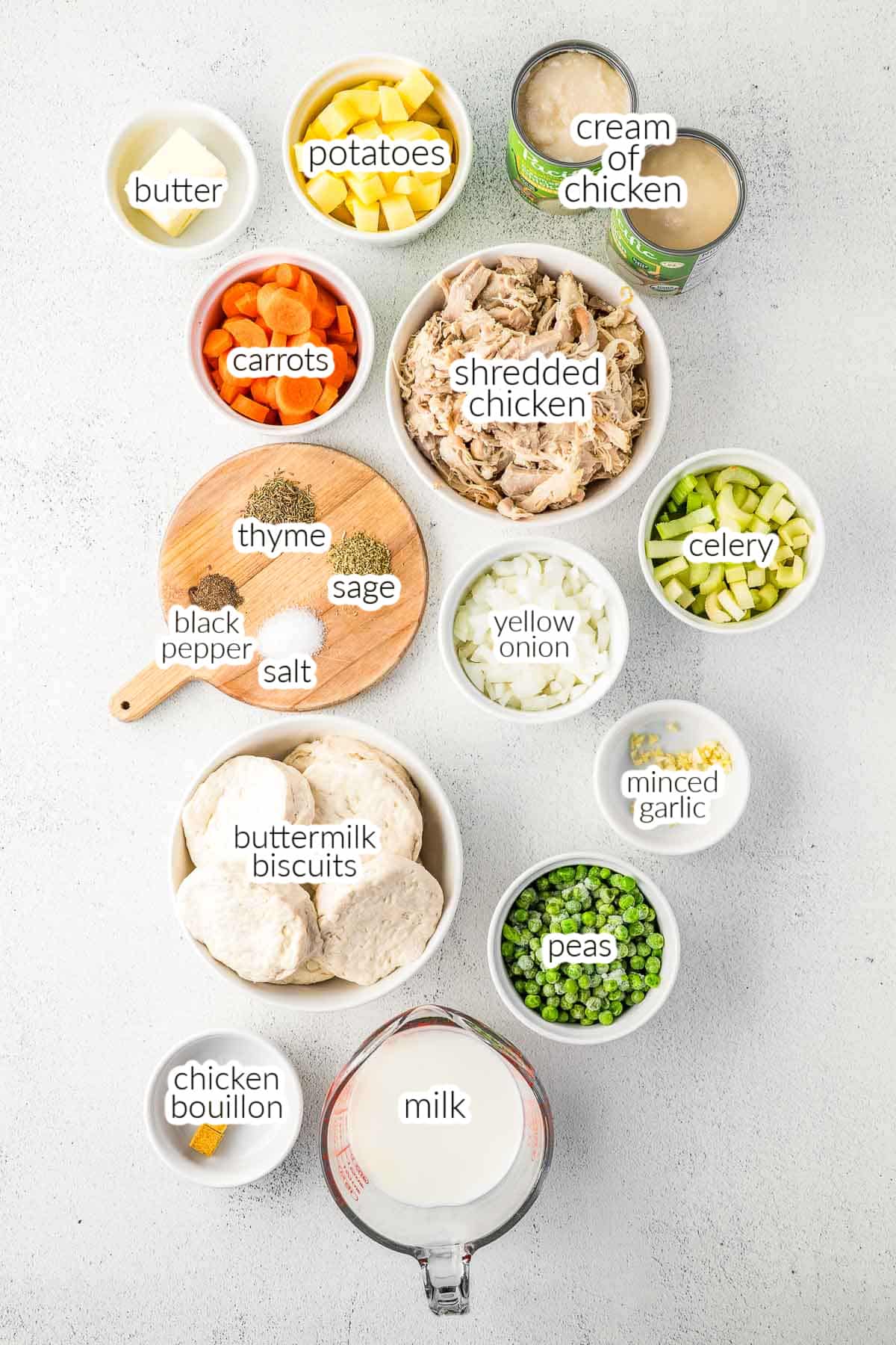 Ingredients for slow cooker chicken pot pie casserole laid out on a white surface, including shredded chicken, vegetables, biscuits, seasonings, cream of chicken soup, milk, and butter.