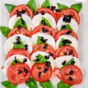 Easy Caprese salad recipe with slices of tomato and mozzarella arranged with basil leaves, topped with balsamic glaze on a white plate.