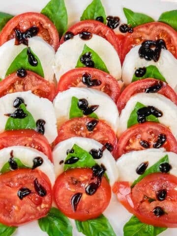 Easy Caprese salad recipe with slices of tomato and mozzarella arranged with basil leaves, topped with balsamic glaze on a white plate.