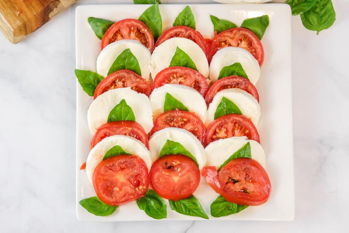 Slices of tomato, mozzarella, and basil leaves arranged in alternating rows on a square white plate.