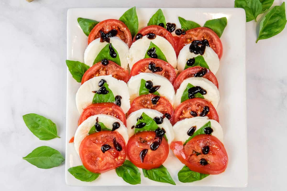 A square plate with rows of sliced tomatoes, mozzarella cheese, and basil leaves, drizzled with balsamic glaze.