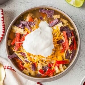 A bowl of chicken tortilla soup topped with shredded cheese, tortilla strips, and a dollop of sour cream.