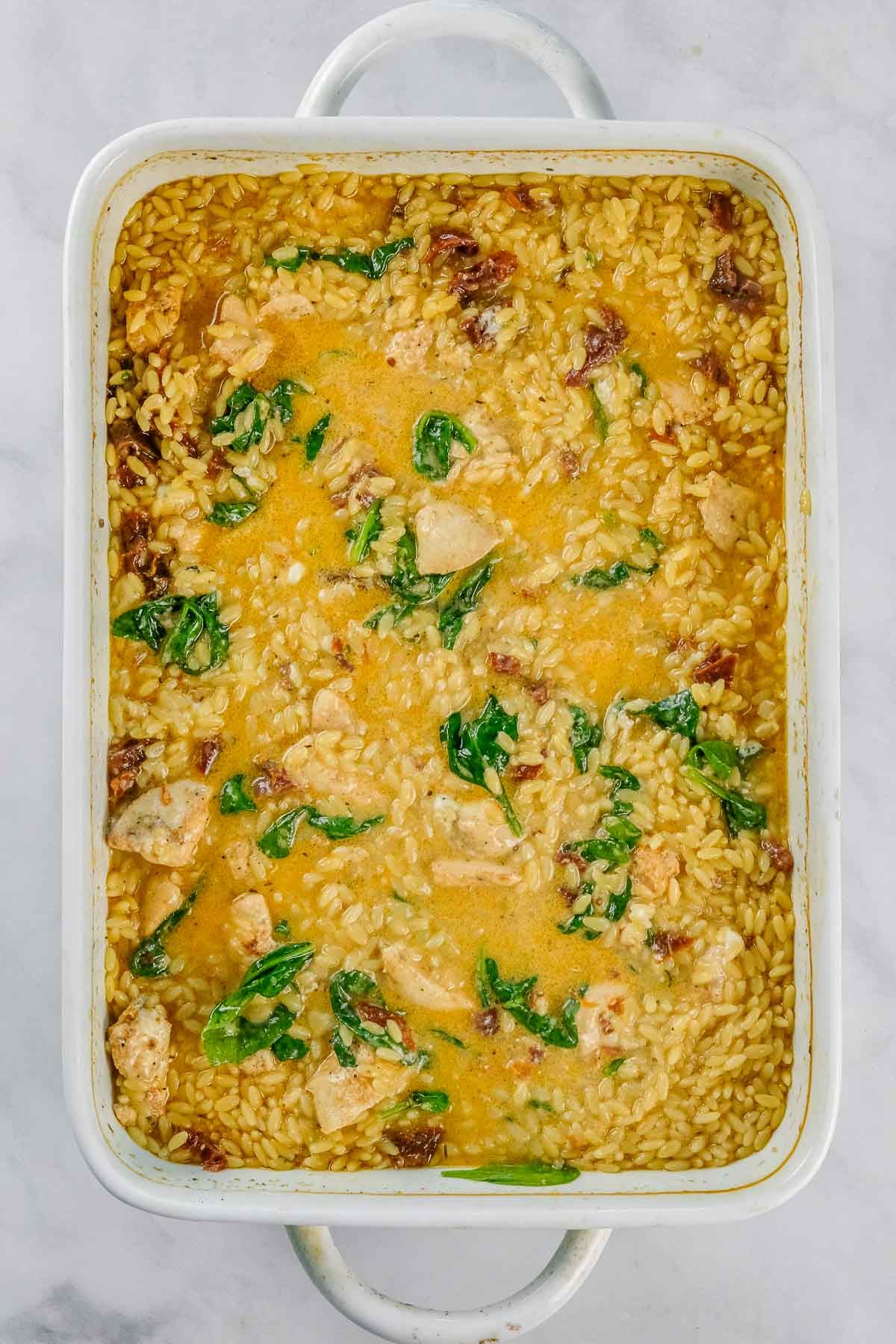 White baking dish filled with Baked Chicken Orzo, spinach leaves, and sun-dried tomatoes.