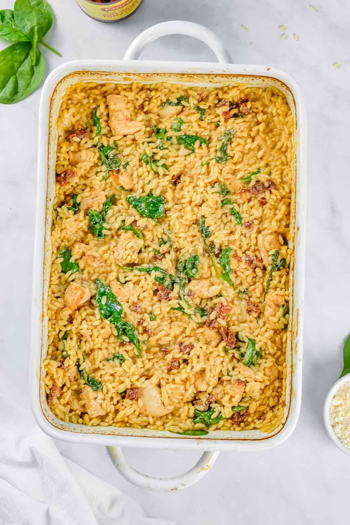A white baking dish filled with Baked Chicken Orzo, spinach, sun-dried tomatoes, and pieces of chicken on a marble surface.