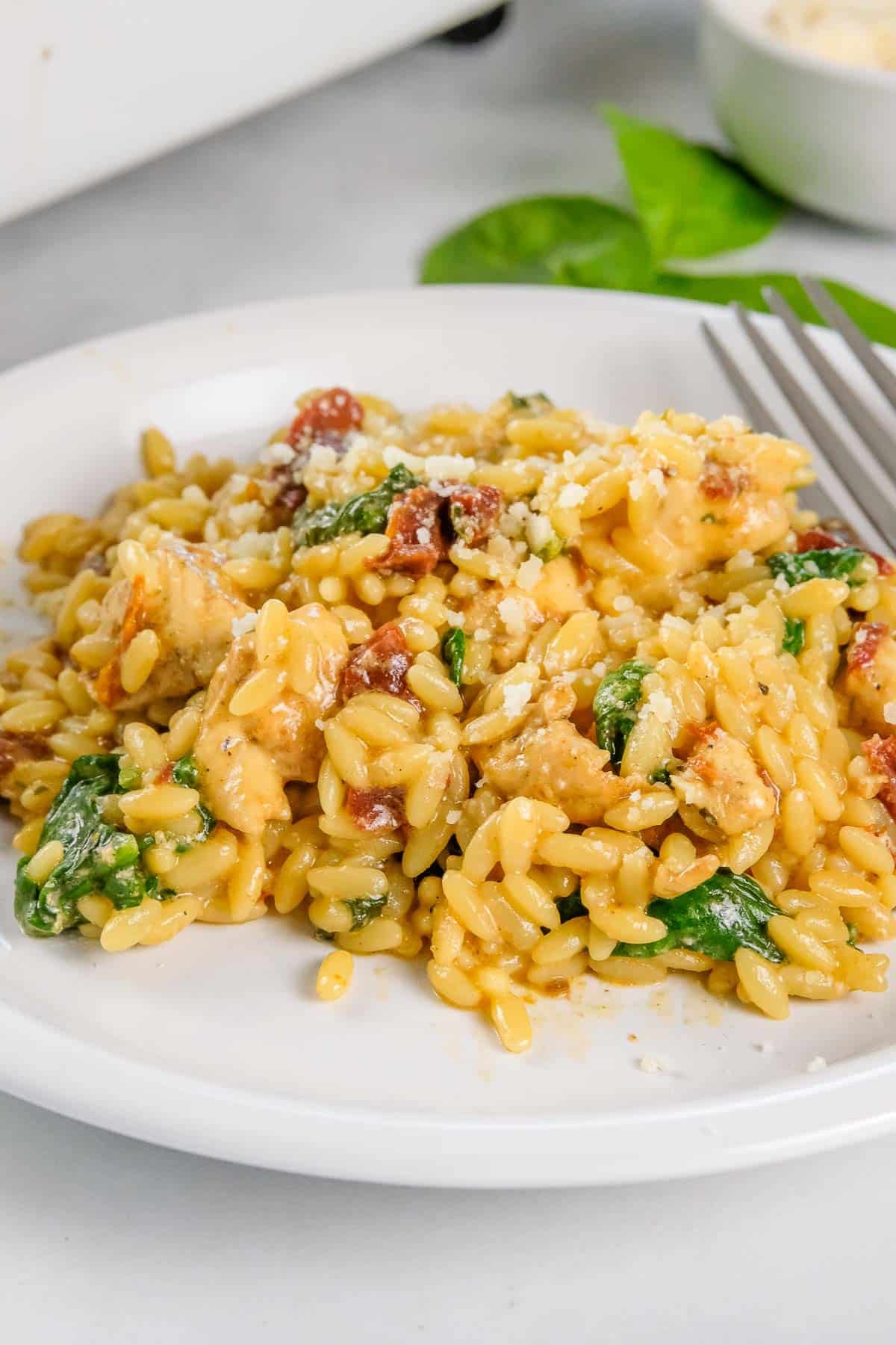 A white plate with baked chicken orzo pasta mixed with spinach, sun-dried tomatoes, and pieces of chicken.