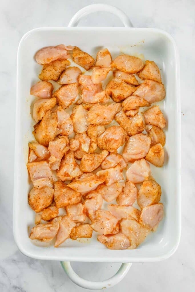 Raw, seasoned chicken pieces arranged in a single layer in a white rectangular baking dish on a marble surface.