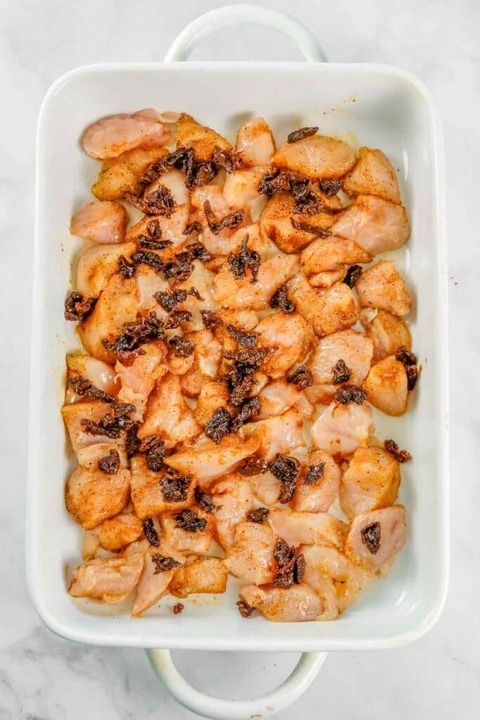 Raw, seasoned chicken pieces topped with chopped sun-dried tomatoes are arranged in a white rectangular baking dish on a marble surface.