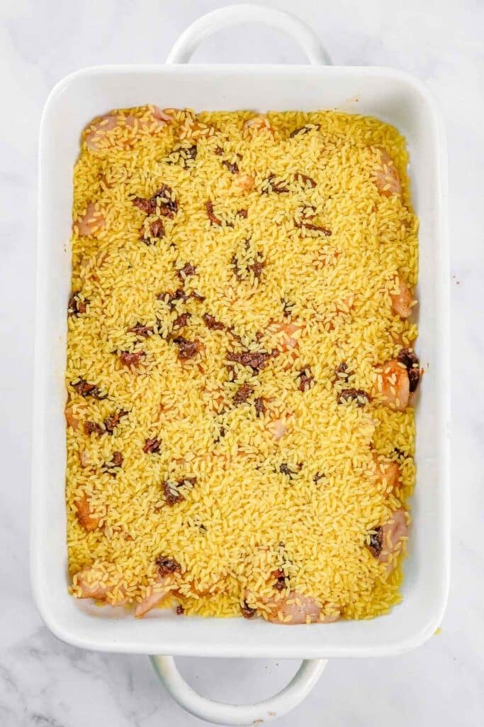 Uncooked orzo, pieces of chicken, and seasoning are spread evenly in a white rectangular baking dish on a marble surface.