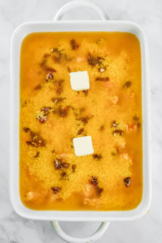 Rectangular white baking dish filled with uncooked oroz, chicken broth, seasonings, and two slices of butter on top.