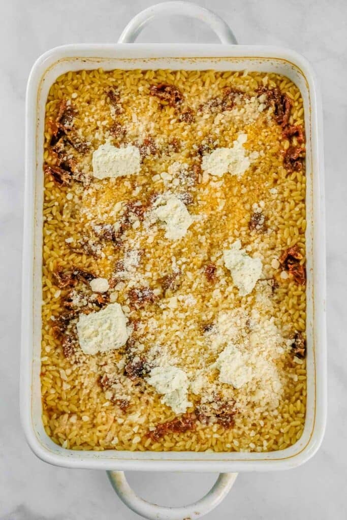 Rectangular white baking dish filled with orzo pasta, sun dried tomatoes, boursin cheese dollops, and grated parmesan on top, set on a light surface.