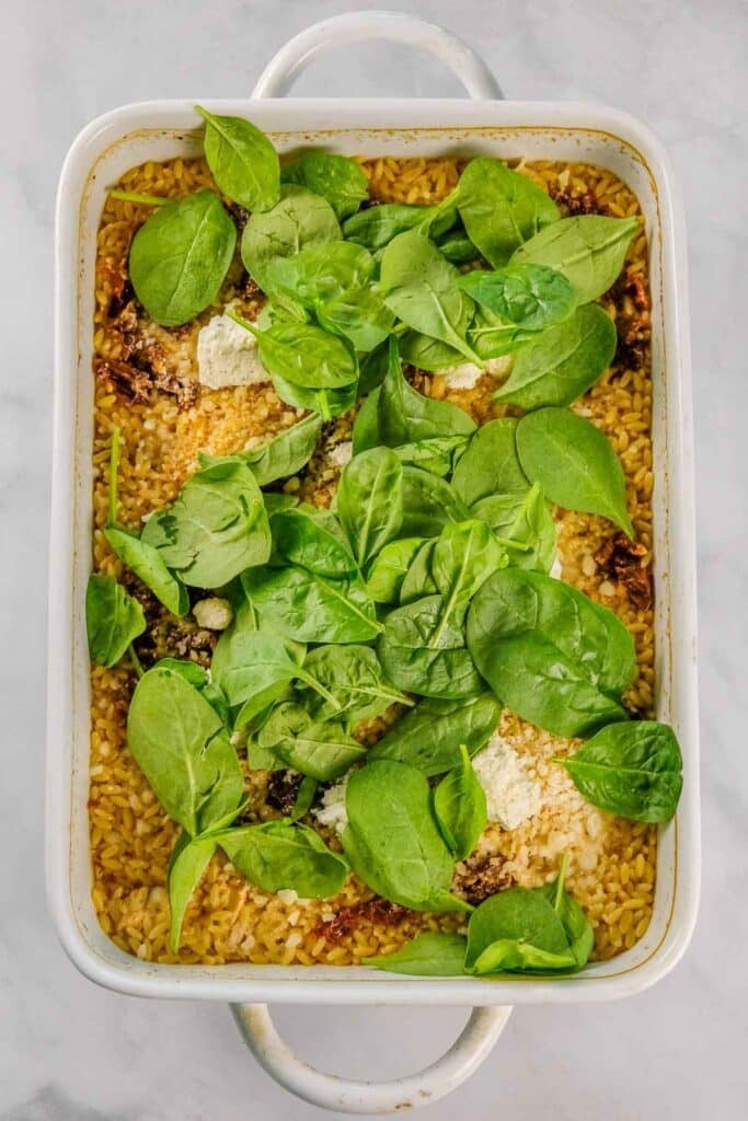 Rectangular white baking dish Baked Chicken Orzo topped with fresh spinach leaves on a marble surface.
