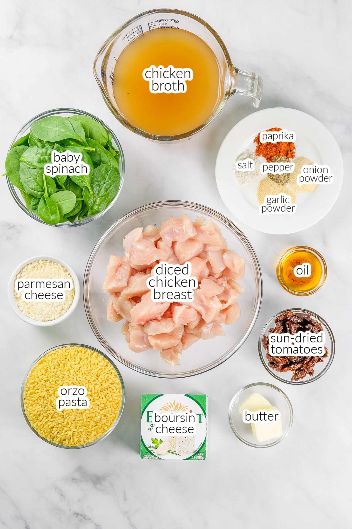Ingredients for a Baked Chicken Orzo dish arranged on a countertop, including diced chicken breast, orzo, spinach, broth, butter, cheese, sun-dried tomatoes, oil, and paprika, salt, pepper, onion powder and garlic powder.