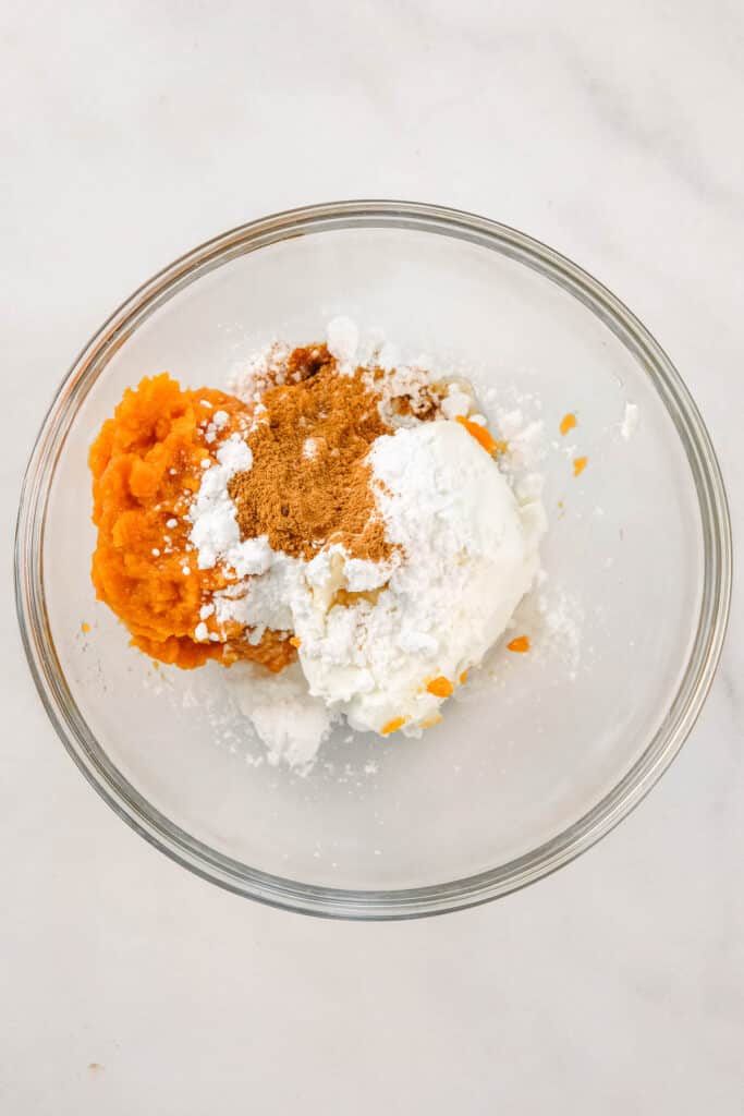 A glass bowl containing pumpkin puree, cream cheese, powdered sugar, and cinnamon on a white surface.