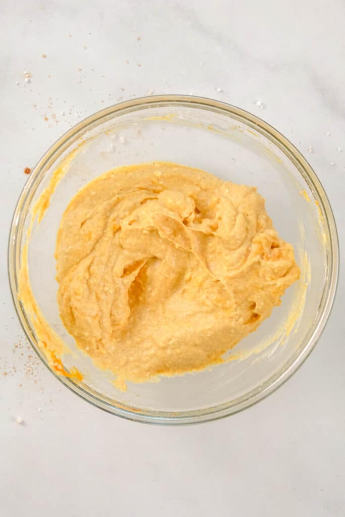 A glass bowl filled with a pumpkin puree batter sits on a white surface with a few crumbs around it.